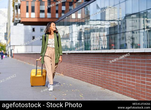 Smiling woman pulling wheeled luggage on footpath