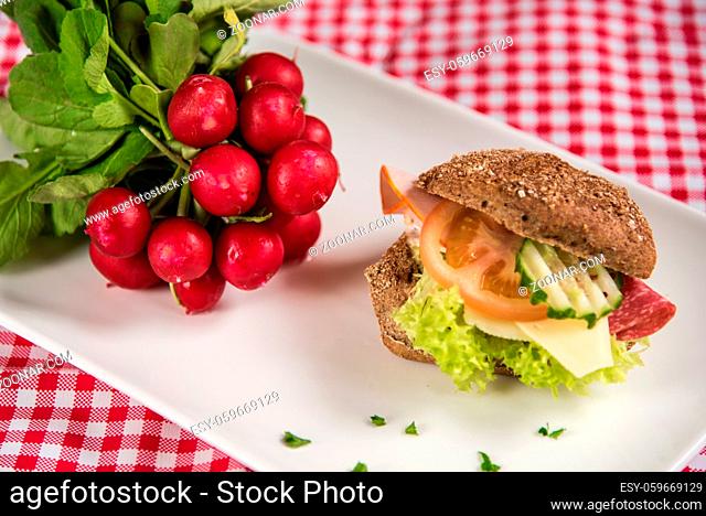 on a white plate lies a sliced bread roll filled with sausage, cheese and egg and as decoration a bunch of radishes
