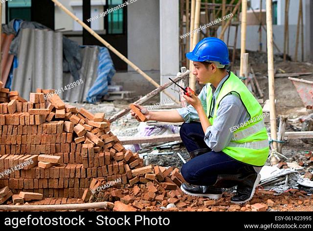 Asian contractor with safety helmet, reflective vest and rubber boots checking quality of brick while holding walkie talkie