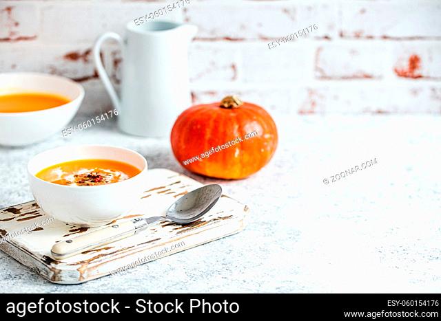 Homemade pumpkin cream soup served in white ceramic bowl on white table with spoon decorated with whole pumpkin, angle view, selective focus