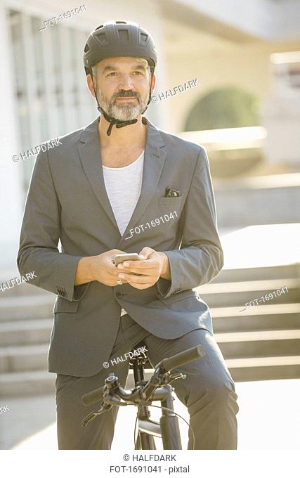 Confident businessman holding mobile phone while sitting on bicycle at city during sunny day