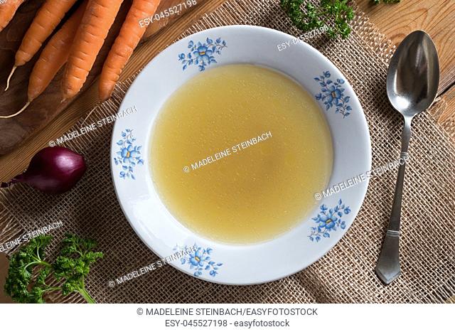 Chicken stock in a white plate, with carrots, onions and parsley in the background, top view