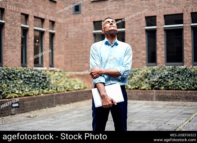 Mature businessman with eyes closed holding laptop in front of office building