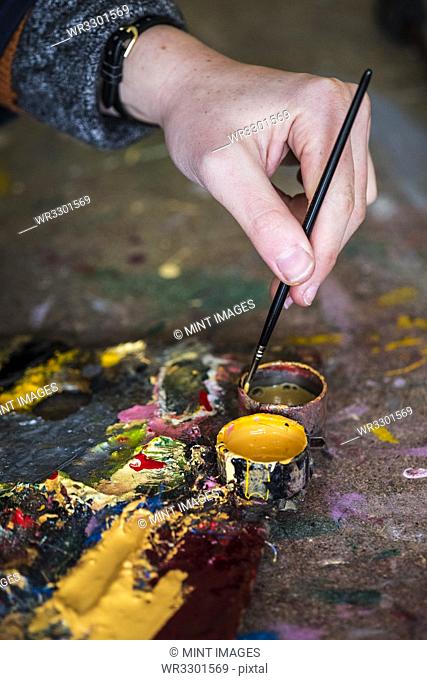 High angle close up of human hand dipping paintbrush into small pot of yellow oil paint