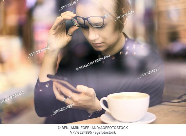 woman behind glass window touching glasses, using smartphone next to coffee cup in café in Munich, Germany