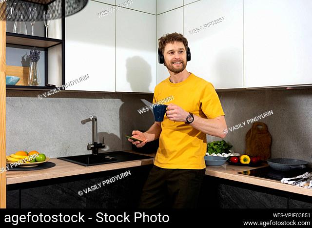 Man listening music while holding mobile phone and coffee cup in kitchen at home