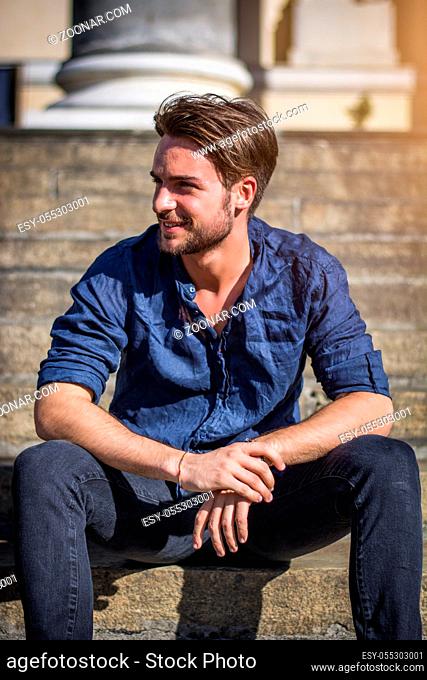 One handsome young man in urban setting in summer day, wearing blue shirt, sitting on stone steps