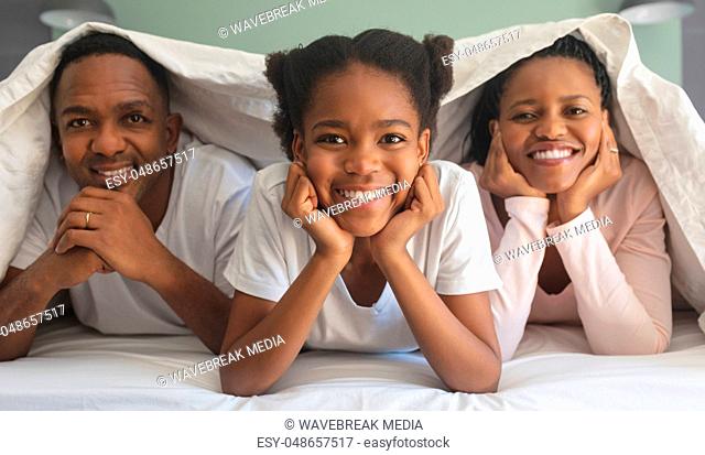 Happy African American family under blanket and looking at camera on bed in bedroom