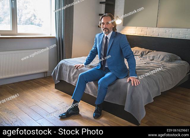 In the bedroom. Man in a blue suit sitting on the bed in the bedroom