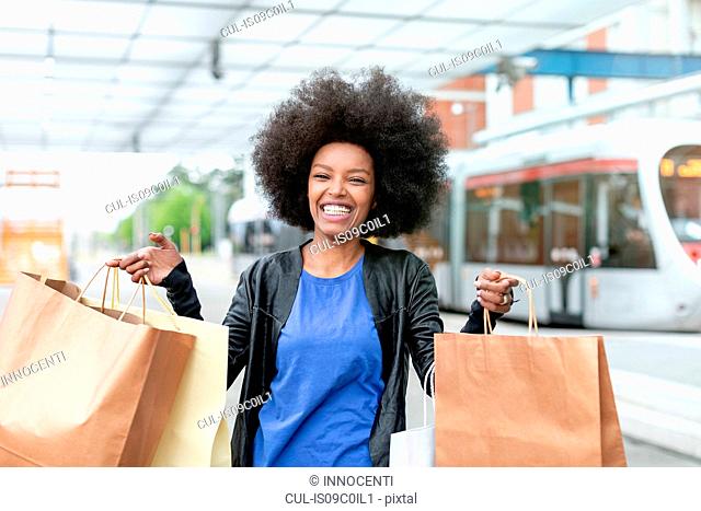 Young woman with afro hair at city train station holding up shopping bags, portrait