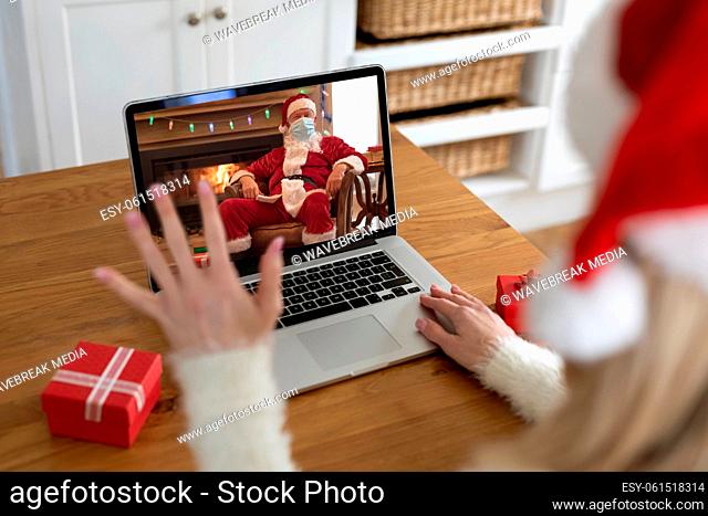 Woman in Santa hat waving while having a video chat with Santa Claus on her laptop at home