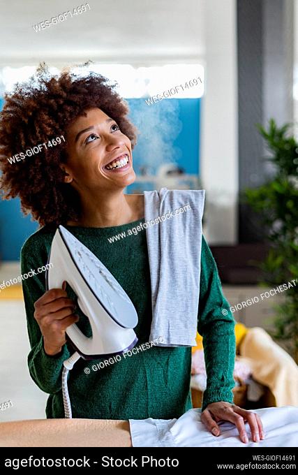 Playful young Afro woman holding iron with shirt on board in living room