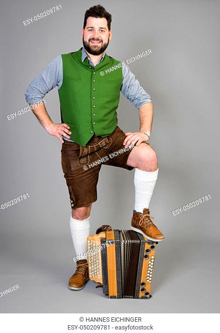 young man with black beard and leather trousers and traditional costume and accordion is posing in front of grey background