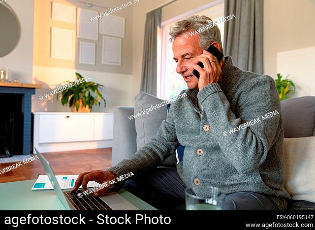 Focused caucasian senior man sitting on sofa, doing paperwork, using laptop and making call