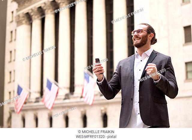 Happy young businessman with cell phone in front of Stock Exchange, New York City, USA