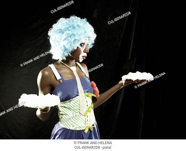 Studio portrait of young woman wearing clown face paint and blue wig holding meringues