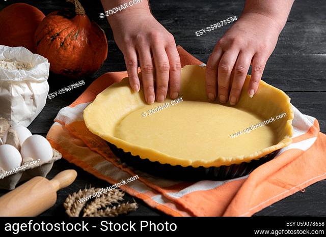 Home baking of holidays classic sweet pies. Woman hands fixing pastry dough in a tray and pumpkin pie ingredients. Preparing traditional desserts