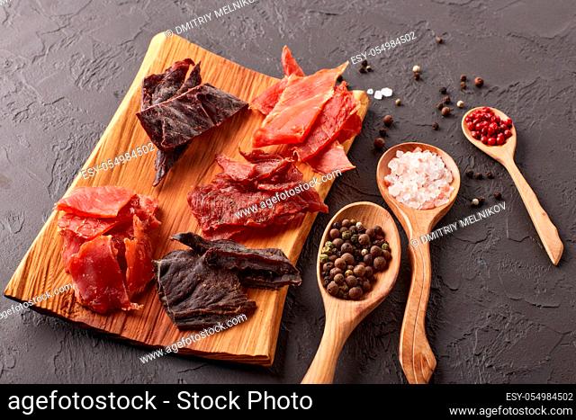 Jerky. Set of various kind of dried spiced meat on wooden tray, diverse peppercorns and salt on wooden spoons on dark gray background. Top view