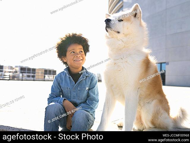Smiling boy looking at dog while sitting on footpath