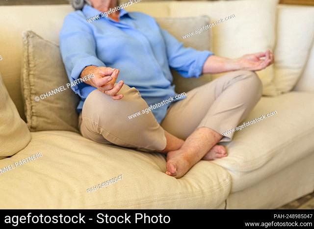 Mid section of senior caucasian woman sitting on sofa meditating. retreat, retirement and happy senior lifestyle concept. || Model approval available