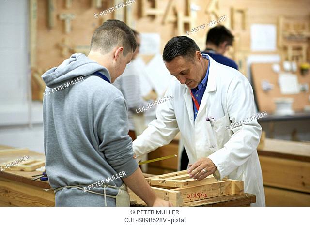 Male lecturer demonstrating measurement to teenage carpentry student in college workshop