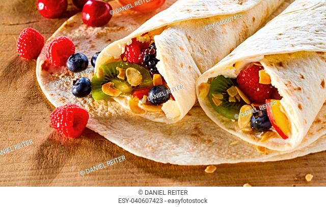 Two tasty healthy summer fruit wraps with roasted mixed nuts and whipped cream on a wooden table with scattered fresh tropical fruit ingredients