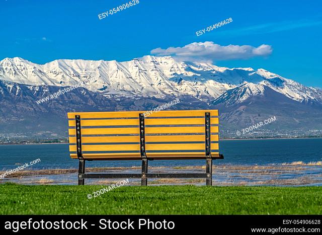 Back view of an empty bench overlooking a calm lake on a sunny day. A snowy mountain against blue sky can also be seen from the outdoor seat