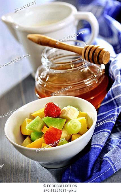 A bowl of fruit salad with a jar of honey and a jug of milk in the background