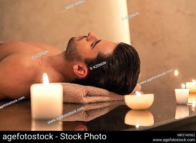 Young man relaxing on massage table surrounded by scented candles at Asian spa and wellness center