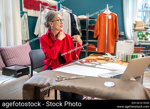 Skill. Inspired gray-haired woman in glasses touching shoulder with measure looking at laptop screen sitting at table in sewing workshop