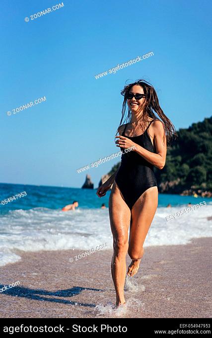 Brunette woman in one-piece swimsuit and sunglasses runs along seashore