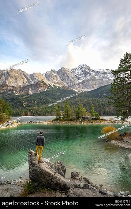 Young man standing on a rock on the shore, view into the distance, Eibsee lake in front of Zugspitze massif with Zugspitze, Wetterstein range, near Grainau