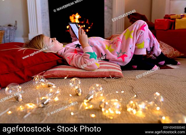 Side view of a happy young Caucasian girl lying on the floor wearing a onesie using a tablet in her sitting room at Christmas time