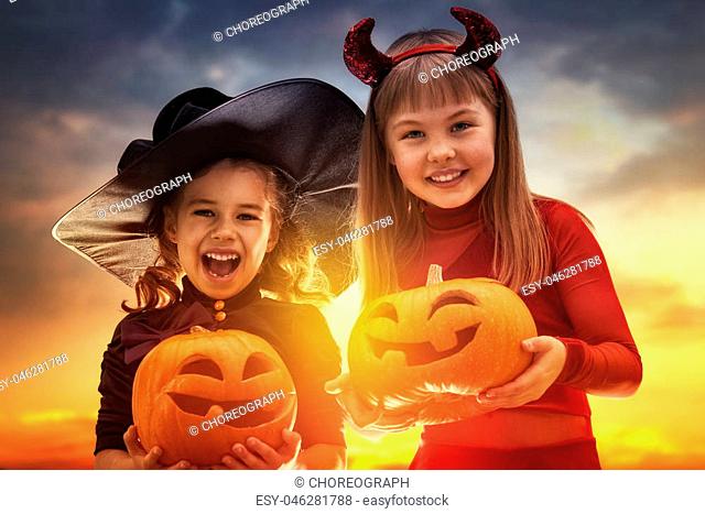 Two happy sisters on Halloween. Funny kids in carnival costumes outdoors. Cheerful children and pumpkins on sunset background