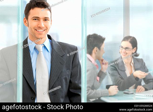 Happy businessman standing in modern glass office, businesswomen sitting at desk in the background, talking