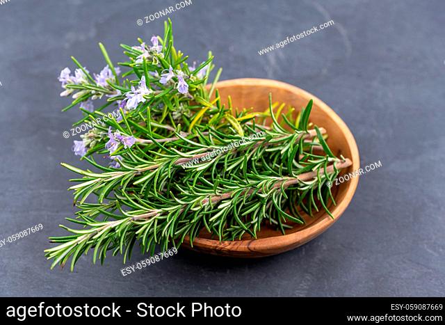 Purple blooming rosemary and fresh rosemary twigs in an olive wood bowl on a black slate background