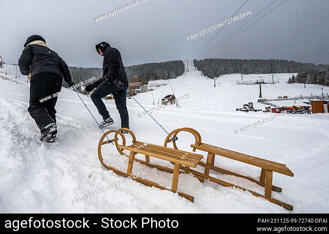 25 November 2023, Saxony, Oberwiesenthal: Two tourists from Saxony-Anhalt go up the ski and toboggan slope on sledges. There was fresh snow in the Erzgebirge...