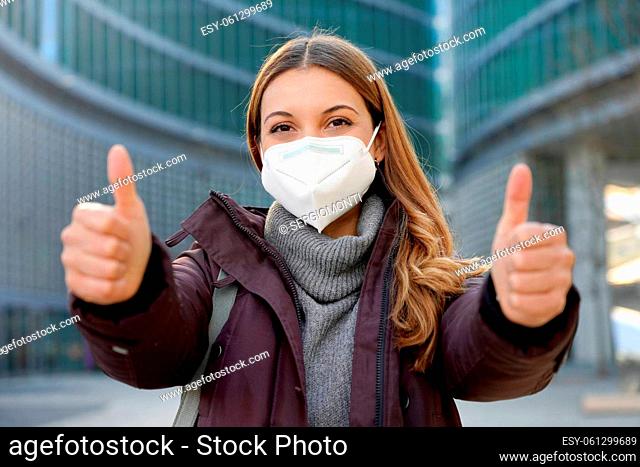 Portrait of optimistic girl wearing protective face mask showing thumbs up in modern city street and looking at camera