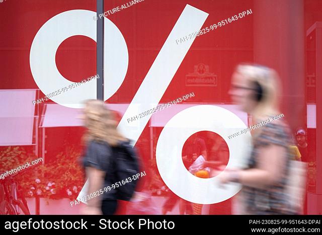 PRODUCTION - 24 August 2023, Saxony, Dresden: Passers-by walk past the window of a fashion store in a pedestrian zone that is advertising a discount promotion