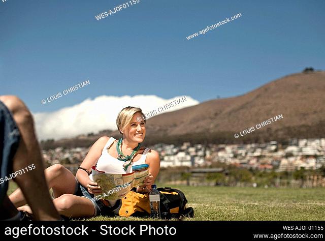 Smiling woman with map looking away while relaxing in public park against sky during weekend