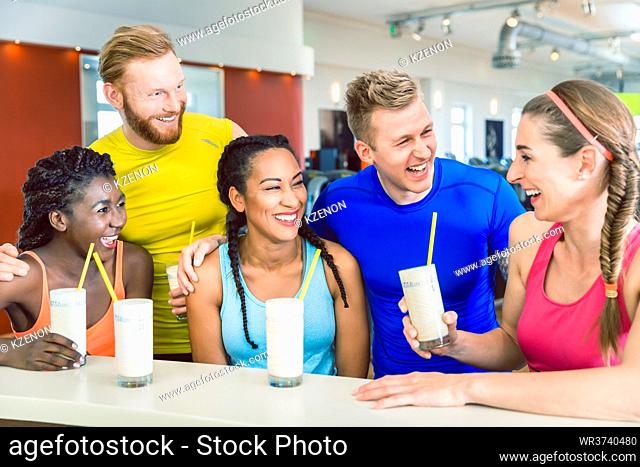 Multi-ethnic group of cheerful friends laughing while drinking white protein shakes before workout at a modern fitness club