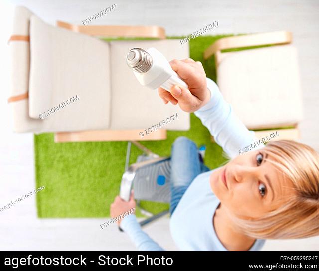 Woman in high angle view trying to change light bulb standing on ladder in living room looking up.Focus placed on light bulb