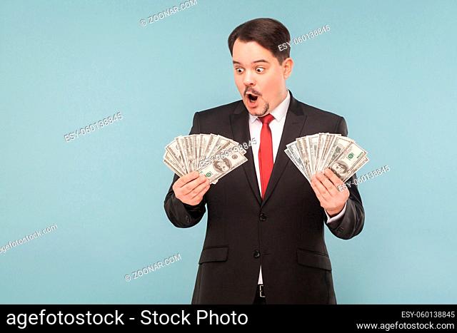 Shocked business man surprised and holding many dollars. Indoor, studio shot, isolated on light blue or gray background