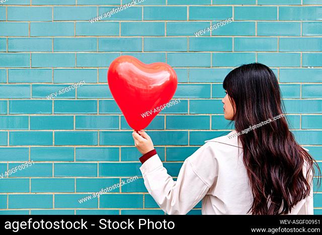Woman looking at heart shape balloon in front of turquoise brick wall