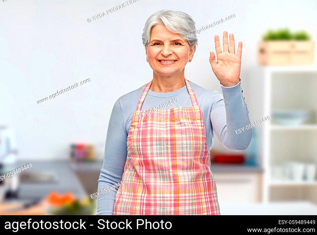smiling senior woman in kitchen apron waving hand