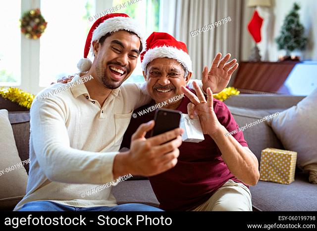 Happy biracial adult son and senior father in santa hats making smartphone christmas video call