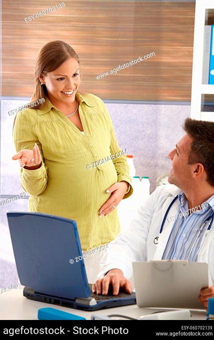 Pregnant woman standing in consulting room, talking with doctor, smiling