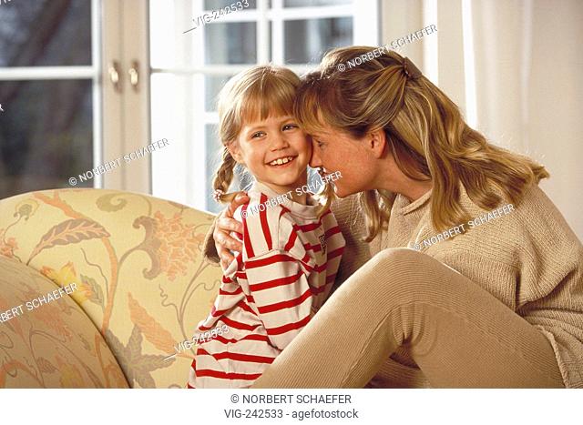 half-figure, indoor, bright dressed blond woman and her 6-year-old blond daughter wearing a red-white striped shirt sit on the sofa smooching  - GERMANY