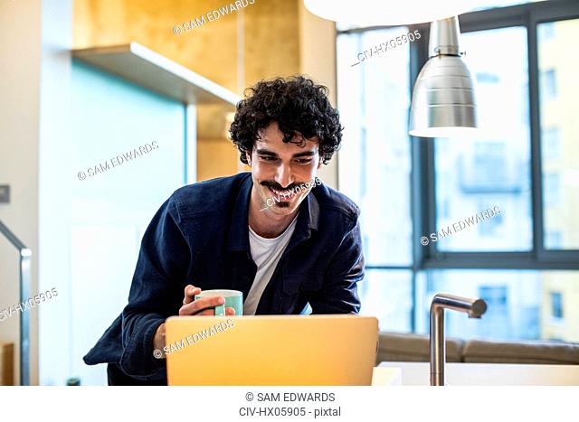 Smiling man drinking coffee, working at laptop in apartment kitchen