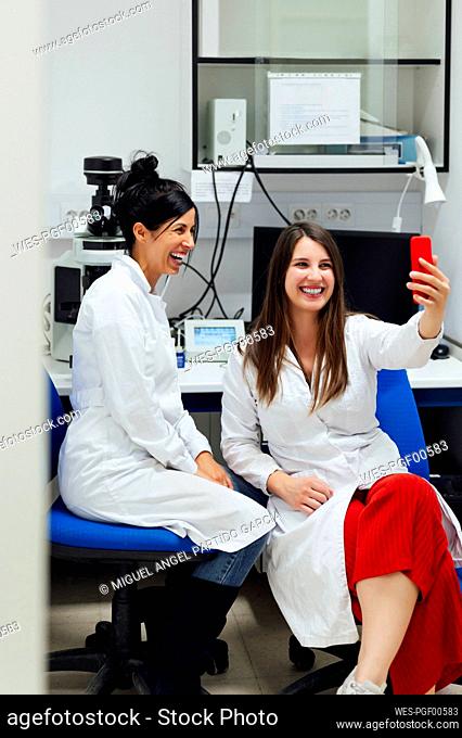 Cheerful young female medical expert taking selfie through smart phone with colleague in laboratory